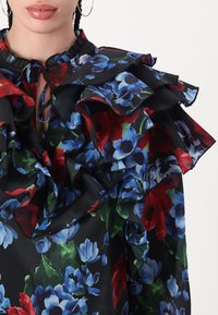 Silk blouse with a black background, featuring colorful floral patterns in red, blue, and green. Ruffled neckline and layered sleeves.