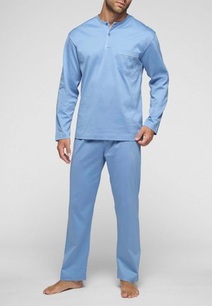 Uomo che indossa un pigiama azzurro chiaro a maniche lunghe con tasca sul petto, in piedi a piedi nudi davanti a uno sfondo chiaro uniforme.
