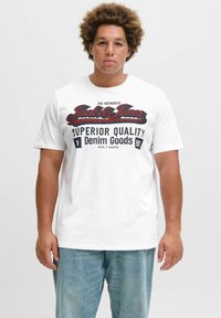 Bijela pametna majica s kratkim rukavima i odvažnim crnim i crvenim grafičkim printom koji glasi "Jack & Jones Superior Quality Denim Goods." Klasičan kroj.