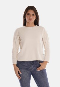 Maglione lavorato a maglia color beige chiaro con scollatura rotonda, maniche lunghe e orlo a coste. Presenta un piccolo logo sul lato. Indossato con jeans blu.