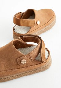 Sandales en suede marron avec une sangle en velcro, doublure intérieure douce et semelle en caoutchouc texturé. Présente un accent de bouton décoratif sur le côté.