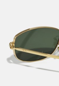 Lunettes de soleil en métal doré avec des verres verts, de forme angulaire surdimensionnée, branches fines et logo gravé sur la monture.