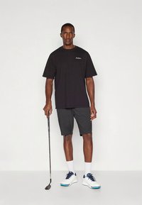 Zwart t-shirt met korte mouwen en logo, gecombineerd met grijze korte broek. Draagt witte schoenen met blauwe accenten en houdt een golfclub vast.