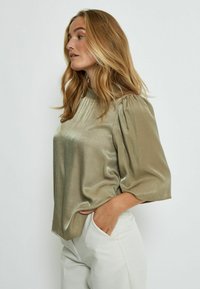 Satin olive green top with puffed sleeves, high neckline, and a relaxed fit. Paired with light-colored pants, soft sheen texture.
