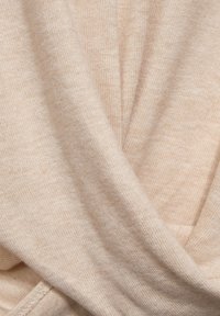 Vaaleanbeige neulottu kangas, jossa on pehmeä tekstuuri. Materiaali laskeutuu pehmeissä laskoksissa, ja siinä näkyy hienovaraisia sävy- ja kudosvaihteluita.