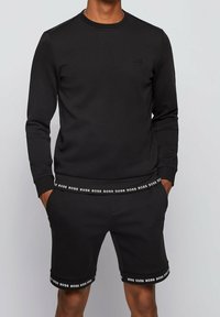 Sudadera negra con cuello redondo y logo bordado, combinada con pantalones cortos a juego que cuentan con una cintura con logo y acentos en el bajo.