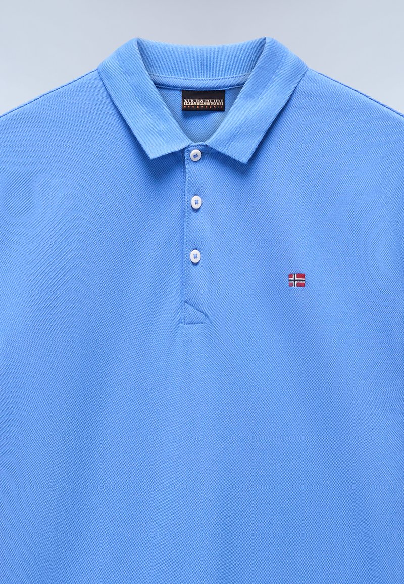 Polo light bleu en coton texturé, avec un col, trois boutons et un petit drapeau norvégien brodé sur la poitrine gauche.