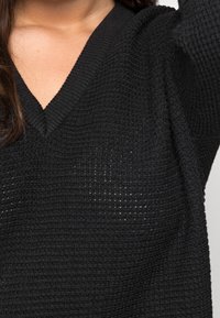 Czarny sweter w serek z teksturowanej dzianiny, charakteryzujący się luźnym krojem i otwartym splotem z widocznymi detalami szycia.