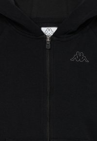 Crna zip-up hoodie s kapuljačom, koja ima mali izvezeni logo dva sjedeća figura leđima okrenuta jedna drugoj na prsima i na povlačenju zatvarača.
