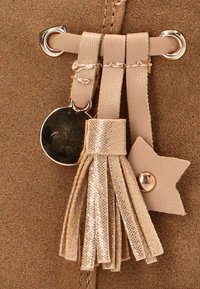 Detalle de un bolso de ante marrón con correas de cuero beige, un charm redondo metálico y un borlón dorado con un pequeño acento en forma de estrella.
