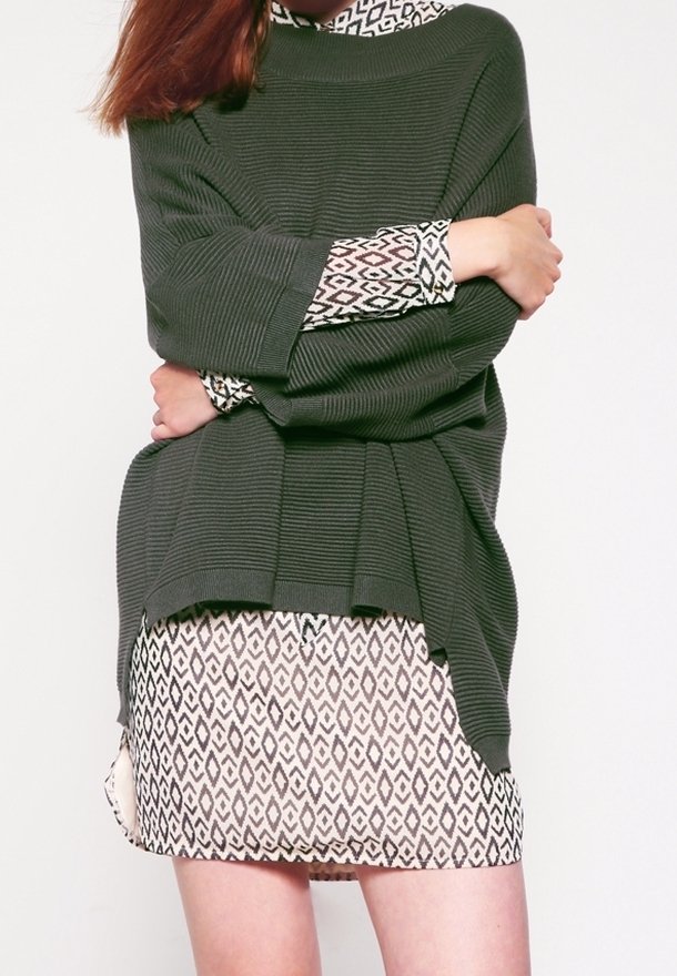 Olivgrön ribbad tröja i stickad kvalitet som bärs över en beige klänning med ett geometriskt svart mönster. Tröjan har breda ärmar och en avslappnad passform.