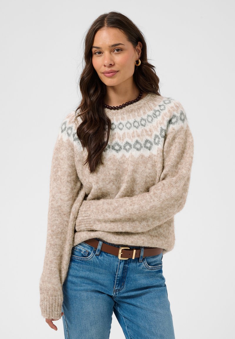 Femme aux longs cheveux foncés portant un pull beige à motifs, un jean bleu, une ceinture marron, des boucles d'oreilles créoles dorées et un collier sombre en perles sur un fond uni.