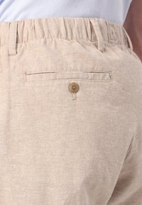 Beige linen shorts featuring an elastic waistband, single back pocket with button closure, and a soft, textured fabric.