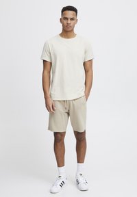 Camiseta de algodón beige de manga corta combinada con pantalones cortos beige. El modelo lleva calcetines blancos y zapatos blancos con rayas negras. Diseño sencillo.
