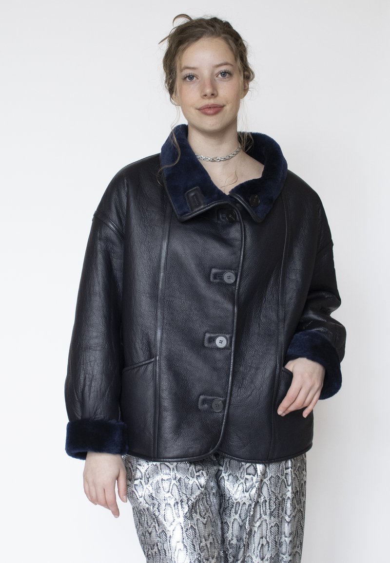 Junge Frau trägt eine schwarze Lederjacke mit dunkelblauem Pelzkragen und -manschetten sowie silberne Hosen mit Schlangenledermuster.