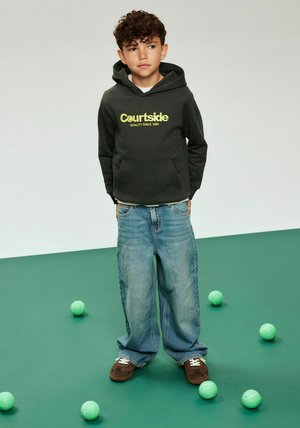 Sudadera con capucha gris oscuro con el logo "Courtside" en amarillo, combinada con pantalones vaqueros azules holgados y zapatillas marrones sobre un suelo verde con pelotas de tenis alrededor.