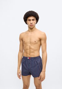 Μπλε ναυτικό μπόξερ με κάθετες κόκκινες ρίγες, ελαστική μέση και λεπτομέρεια με κουμπί στο μπροστινό μέρος. Δεν φαίνονται επιπρόσθετα υλικά ή σχέδια.