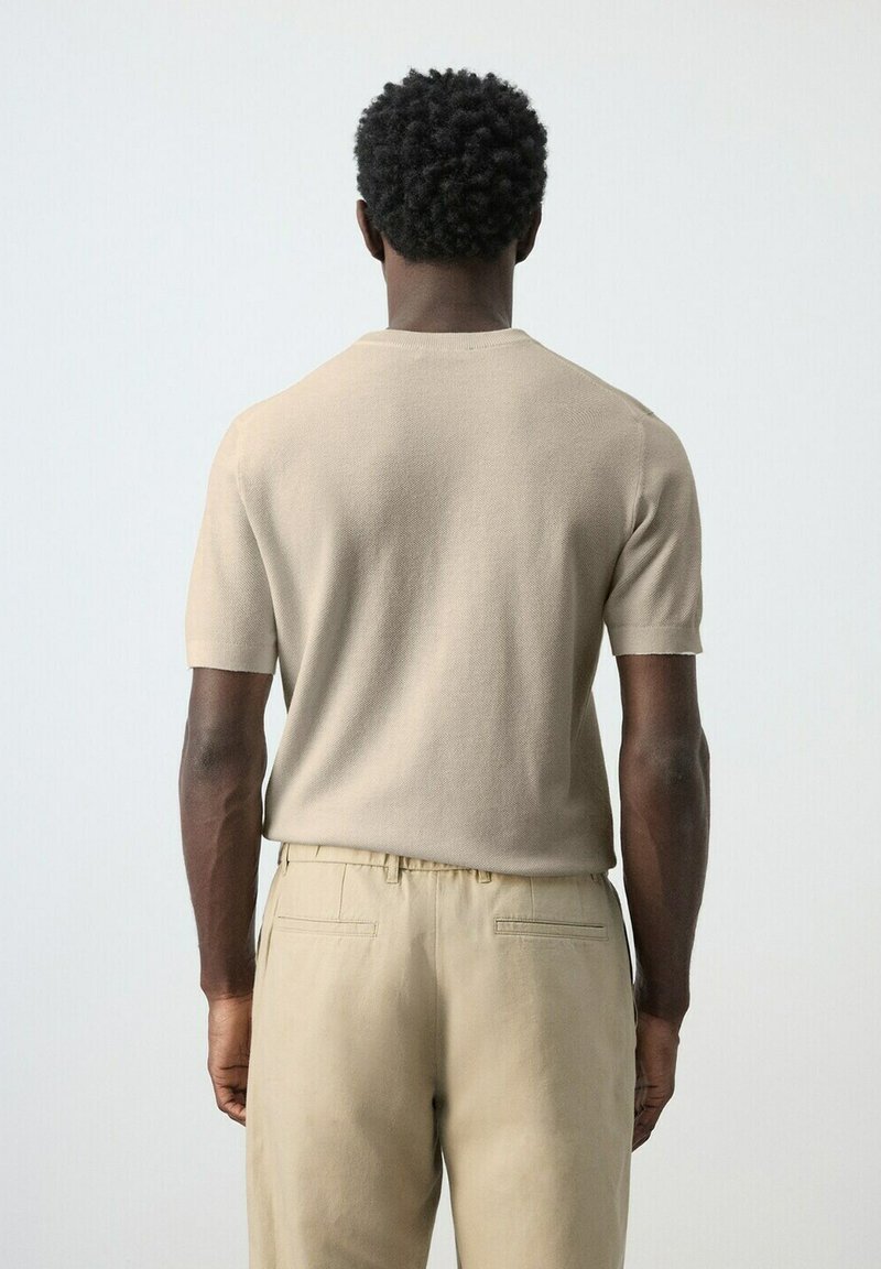 Homme aux cheveux courts et bouclés vu de dos, portant un haut en maille beige à manches courtes et un pantalon beige, sur un fond uni clair.