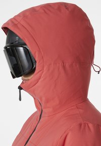 Rote isolierte Jacke mit Kapuze, die einen hohen Kragen, einen schwarzen Reißverschluss und ein strukturiertes Material aufweist. Sie hat eine taillierte Passform und eine Schnürung an der Kapuze.
