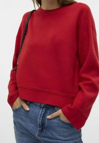 Sweat-shirt rouge en coton avec col et poignets côtelés, doté d'un ourlet à bandes et d'une coupe décontractée, associé à un jean en denim bleu.