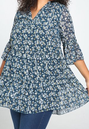 Kvinde iført marineblå blomstret tunika med 3/4 rynkede ærmer og lagdelt kant, kombineret med mørkeblå jeans.