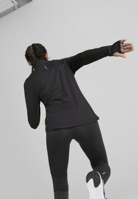 Chaqueta deportiva negra con cuello alto, mangas largas y agujeros para los pulgares. Combinada con leggings negros ajustados. Textura suave y diseño minimalista.