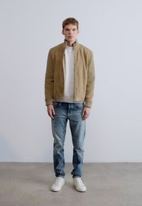 Világosbarna szarvasbőr kabát, bordás fehér pulóver, világoskék farmer és fehér sportcipő. Betonpadlón áll, egyszínű háttér előtt.