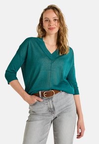Un pull en tricot turquoise, ample, avec un col en V et des manches trois-quarts, associé à un jean gris clair et une ceinture marron avec une boucle dorée.