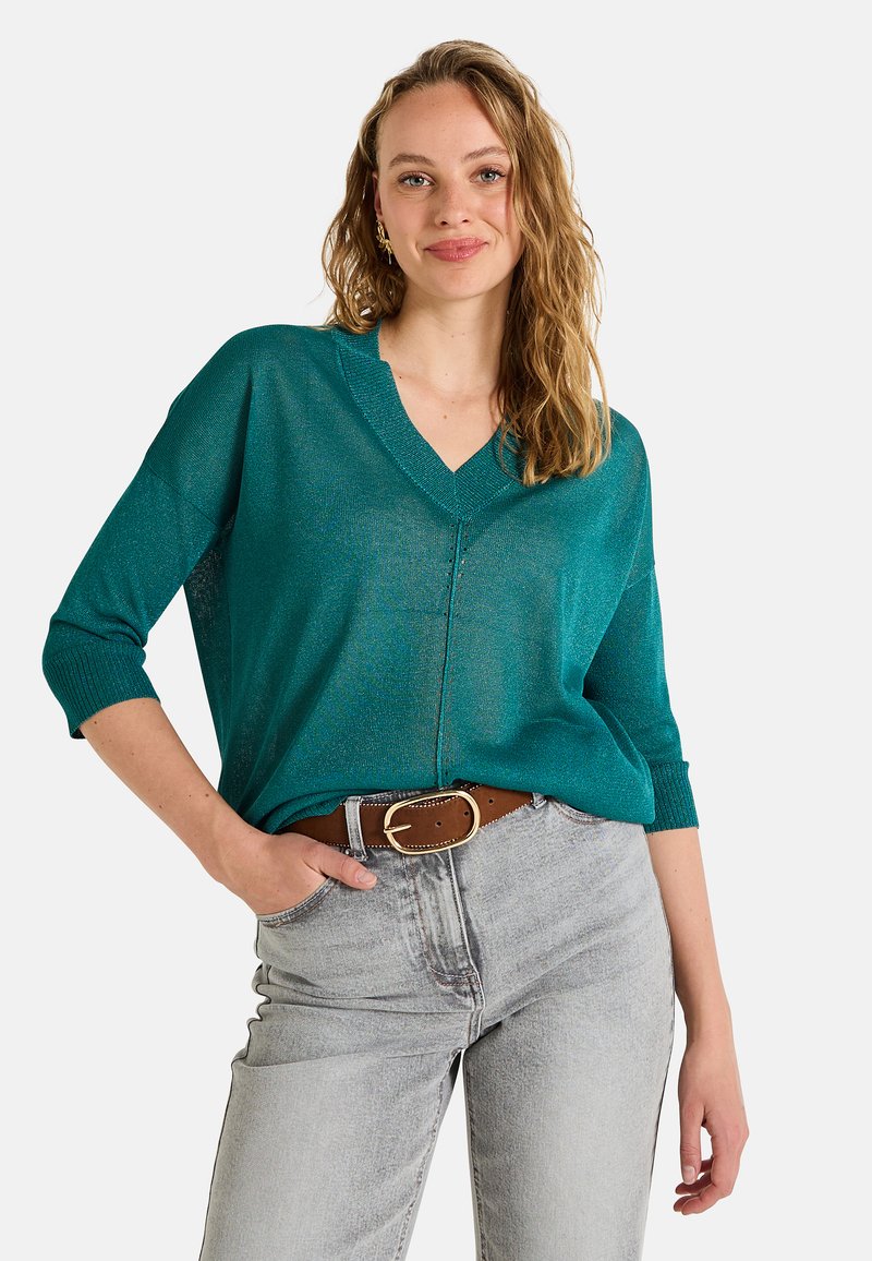 Un pull en tricot turquoise, ample, avec un col en V et des manches trois-quarts, associé à un jean gris clair et une ceinture marron avec une boucle dorée.