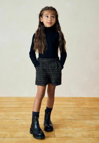 Col roulé noir à côtes, short en tweed avec poches et bottines noires zippées. Fond en bois avec un mur neutre.