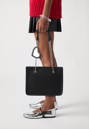 Persona con suéter rojo y falda de cuadros sostiene un bolso negro con un colgante en forma de corazón, combinado con zapatos planos plateados metálicos sobre un fondo blanco.