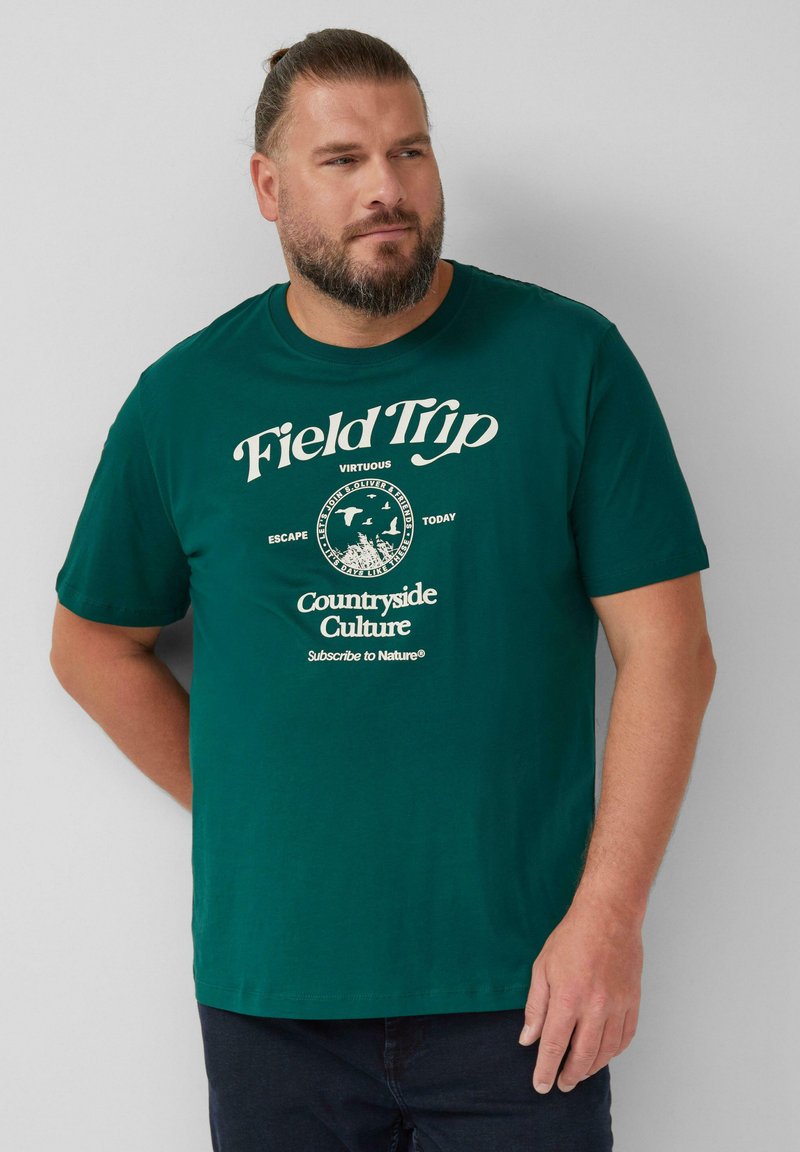 Man met baard in een groen T-shirt met de tekst "Veldtrip Platteland Cultuur" en een natuurafbeelding, staande tegen een witte muur.