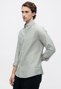 Jovem com cabelo ondulado, vestindo uma camisa de botão cinza claro e calças pretas, de pé com uma mão atrás das costas.