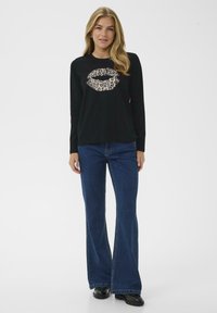 Cămașă neagră cu mânecă lungă, cu un grafic al unui ruj în imprimeu de leopard, asociată cu blugi evazați albaștri și pantofi negri. Look simplu și casual.
