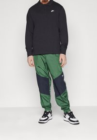 Mężczyzna w czarnej, długosleeve'owej koszuli polo Nike, zielono-czarnych spodniach dresowych Nike i czarno-białych butach Nike stoi na tle jednolitym.