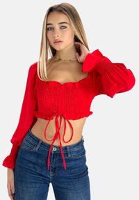 Top corto rojo con escote cuadrado, cuerpo fruncido y mangas largas abullonadas. Se ata a la cintura y tiene un detalle de volante en el dobladillo. Combinado con vaqueros azules.