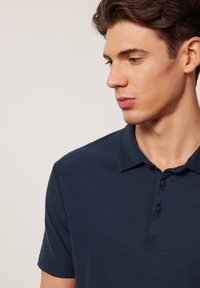 Marinblå polo skjorta med en mjuk textur, korta ärmar och krage. Har tre svarta knappar vid halsen och en avslappnad passform.