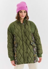 Prešívaná zelená bunda s pletenými detailmi a vreckami s kvetinovým vzorom. Má vysoký golier a fialové patentné gombíky. Nosená ružová čiapka.