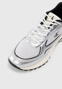 Baltas sportinis batelis su sidabro akcentais, tinklinė tekstūra, juodi akcentai ir balti raišteliai. Yra logotipas ir paminkštintas padas.