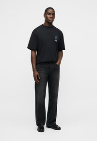 Άνδρας όρθιος, φορώντας μαύρο μπλουζάκι με στάμπα, σκουρόχρωμα γκρι τζιν και μαύρα παπούτσια, με ένα χέρι στην τσέπη, μπροστά σε ένα απλό φόντο.