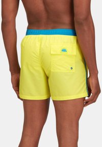 Bañador amarillo con una cinturilla elástica azul, franjas laterales y un bolsillo trasero con un logotipo azul acentuado. Material suave y ligero.