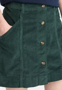 Groene corduroy rok met vijf messing knopen en een grote zijzak. Heeft een verticale ribbelstructuur, rechte snit en een ruwe zoom.