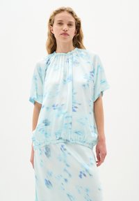 Ljusblå blus med kort ärm i ett flödande, genomskinligt tyg med en samlad halsringning och elastisk nederkant som har ett subtilt vatt färgsmönster.