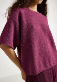 Maglione lavorato a maglia viola con maniche corte, caratterizzato da una texture morbida e pelosa e una vestibilità comoda, presentato su uno sfondo chiaro.