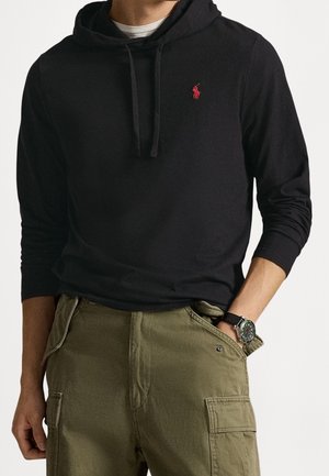 Mann trägt schwarzen Hoodie mit rotem Logo und olivgrünen Cargohosen, Hand in der Tasche, Armbanduhr am linken Handgelenk.