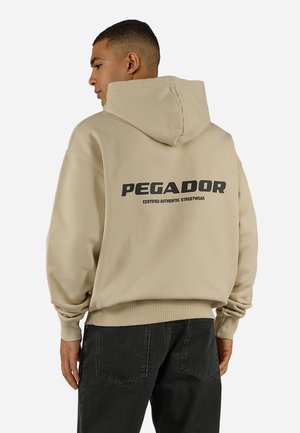 Mężczyzna w beżowej bluzie z kapturem opuszczonym, na plecach czarny napis „PEGADOR Certified Authentic Streetwear”, do tego czarne spodnie.