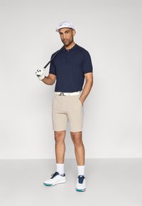 Ναυτικό πόλο μπλούζα, μπεζ σορτς, άσπρη ζώνη και άσπρα παπούτσια γκολφ με μπλε λεπτομέρειες. Κρατώντας μια ρακέτα γκολφ, φορώντας ένα άσπρο καπέλο και γάντια.