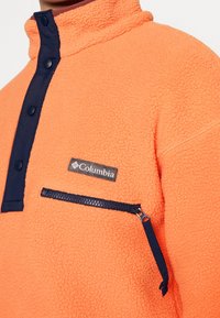 Primer plano de un pulóver de forro polar naranja con botones de presión negros, un bolsillo con cremallera negra y un parche con el logo de Columbia en el pecho.