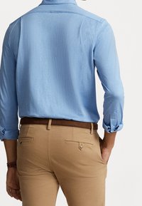 Jasnoniebieska koszula z długim rękawem z podwiniętymi mankietami, zestawiona z beżowymi spodniami z brązowym paskiem i tylnymi kieszeniami zapinanymi na guziki.