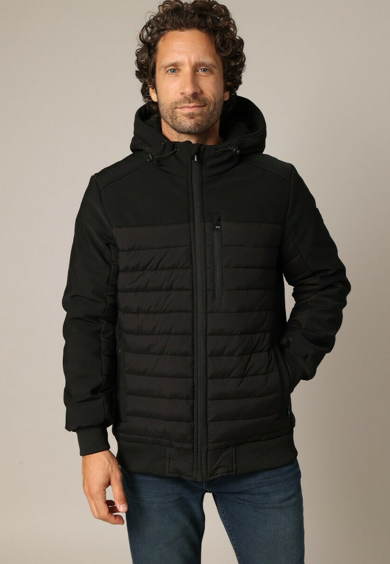 Veste noire avec un devant rembourré et un dos uni, poche poitrine zippée, capuche ajustable et poignets côtelés. Portée sur un jean bleu.